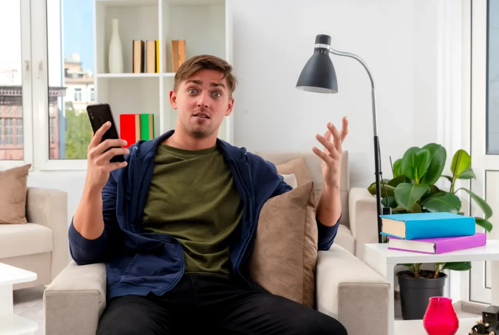 Ung man som ser tveksam och förvirrad ut med sin smartphone i en soffa, illustrerar beslutsångest mellan IPTV månads- eller årsabonnemang.